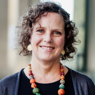 A smiling woman with curly hair, wearing a black top and colorful beaded necklace, stands in a bright outdoor setting.