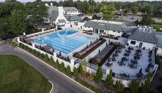A spacious pool area features multiple lanes, surrounded by lounge chairs and greenery, with dining spaces nearby and a charming building in the background.