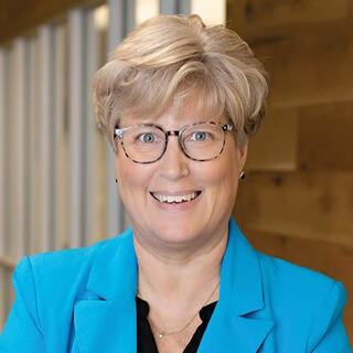 A smiling woman with short, light blonde hair wears glasses and a bright blue blazer, standing in a well-lit, modern setting.