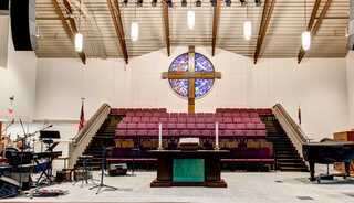 A spacious worship space features a large stained glass window, a cross, tiered seating, and a stage with musical instruments.