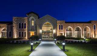 A beautifully lit stone worship building at dusk, featuring arched windows and a landscaped pathway leading to the entrance.