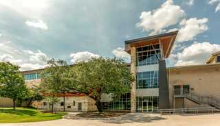 A children's ministry building features large windows and a unique roof design, surrounded by greenery and a clear blue sky with fluffy clouds.