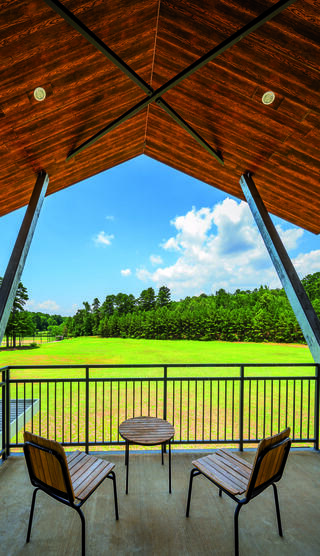 A spacious balcony features two chairs and a table, overlooking a serene green landscape under a bright blue sky with fluffy clouds.