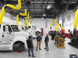 Three mechanics discuss work in a spacious shop with multiple trucks and yellow exhaust hoses hung from the ceiling. Tools and equipment are visible.