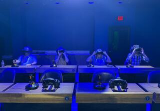 The new Dreamscape Learning VR classroom at the Center for Immersive Learning at St. Charles Community College, with blue lighting, features four people sitting at desks wearing virtual reality headsets