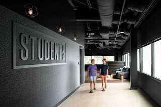 Two young students walk down a hall that has windows on one side and a dark, moody graphic wall with a large sign that says STUDENTS along the corridor and three modern lantern lights hanging from the ceiling. The design is very minimal with black paint and graphics, a light flooring and a couch in the background.