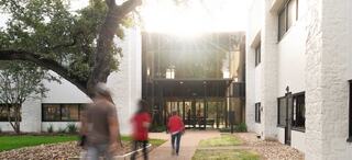 Sundrenched image of a handful of people walking into a white building with black trim windows at Austin Stone West campus in Texas; light from the sun pours through the trees, evoking a glowing type of atmosphere.