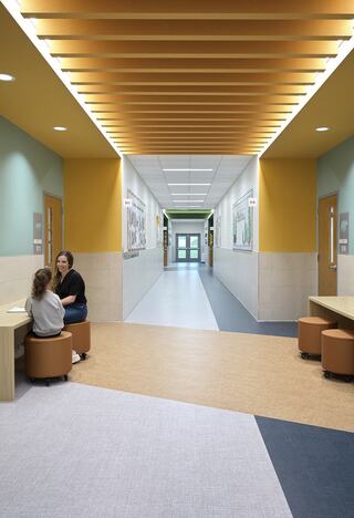 A spacious hallway with colorful walls, wooden ceilings, and seating arrangements. Two people converse near a table, creating a welcoming atmosphere.