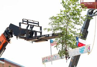 Crane raising signed white steel beam into the air and into place atop the highest point of the building frame at the Houston Elementary School topping out ceremony. A small sapling tree is attached to the middle of the beam and is sandwiched between an American flag and Texas flag