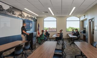 A presenter speaks to a small group seated in a modern classroom, featuring large windows and tables arranged for collaboration.
