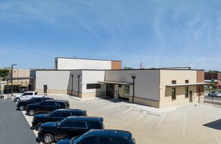 A modern, two-story building with a light exterior and parking area filled with black SUVs under a clear blue sky.
