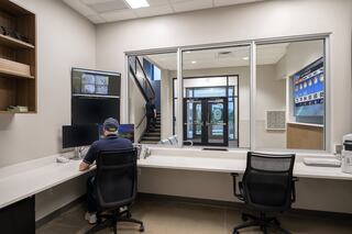 A modern office space features a desk with dual monitors, a view of an entrance, and a person monitoring security footage.