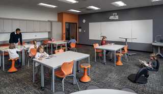 A modern classroom features engaged students at flexible desks that have various seating options, with a teacher assisting. Bright colors and unique seating enhance the learning environment.