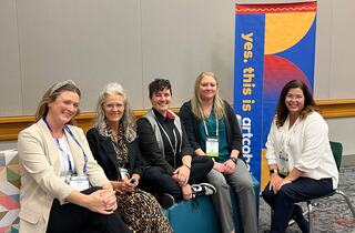 Five women sit together, smiling, in a conference setting, with a colorful banner in the background promoting art education.