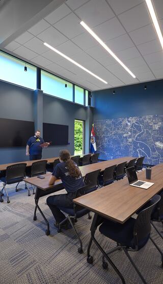 A modern meeting room features a long table, chairs, large screens, and a map mural, with one person presenting to a seated individual.