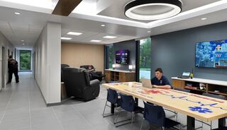 A modern lounge featuring a large table, comfortable seating, a TV, and individuals engaged in leisure activities. Bright, spacious design.