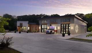 A modern emergency services building features large windows, an ambulance at the entrance, and a landscaped exterior in a serene setting.