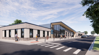 A modern building with a glass entrance and stone facade, featuring people walking nearby and cars parked along the street. Bright and open sky.