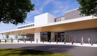 A modern school building with a sleek entry design features large glass entrances, a wooden overhang, and green landscaping, attracting visitors outside.
