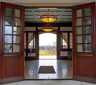 Double wooden doors open to a spacious hallway with elegant lighting, leading to a view of trees and greenery outside.