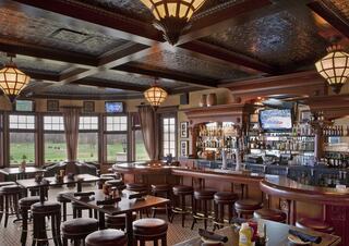 A cozy pub interior features a bar with stools, warm lighting, and large windows offering views of a lush green landscape.
