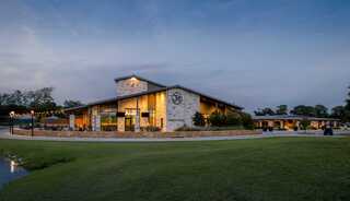 A spacious, elegant home sits by a calm body of water, surrounded by lush green lawns and trees under a soft twilight sky.