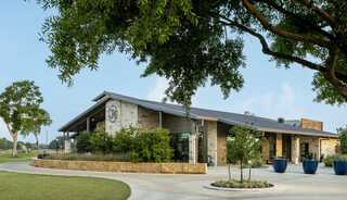 A modern stone building with a sleek roof, surrounded by green grass and trees, features a welcoming entrance and large planters.