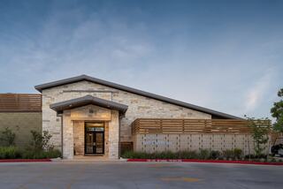 A modern golf clubhouse featuring a stone exterior, wooden accents, and landscaped surroundings, set against a clear blue sky.