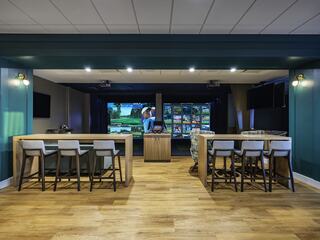 A sleek indoor space features a bar area with high stools, wooden floors, and a golf simulator screen in the background. Modern and inviting.