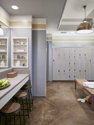 A modern locker room features sleek lockers, a sitting area with bar stools, and stylish decor in soft colors. Games and fruit are present.