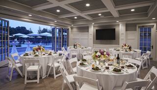 Elegant dining setup with round tables, white tablecloths, and floral centerpieces, overlooking a pool area at dusk.