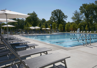A serene outdoor swimming pool is surrounded by sun loungers and umbrellas, set against lush greenery and a clear blue sky.
