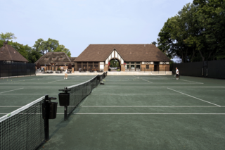 A spacious tennis court area with a charming clubhouse in the background, surrounded by greenery and players practicing.