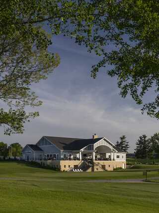 A charming two-story building with a large porch sits amidst lush greenery, framed by trees and a clear sky.