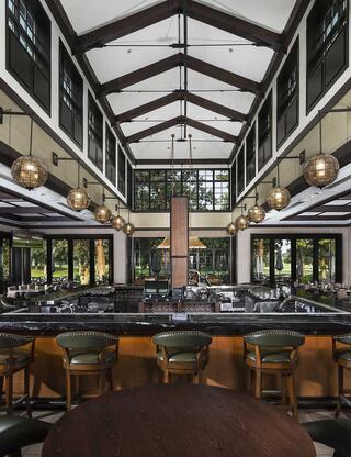 A spacious, elegant restaurant with high ceilings, wooden beams, and a stylish bar area, featuring ambient lighting and large windows.