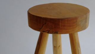 A wooden stool features a round top with visible grain and slight imperfections, supported by three sturdy legs. Minimalistic design.