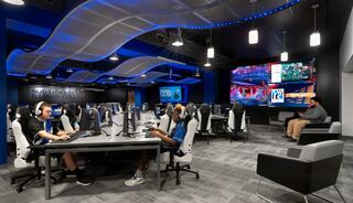 Southwestern Illinois College Esports arena with two students on computers in the foreground and another student in the background; large screen in back, blue LED lighting and modern computer station setup.