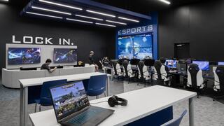 St. Louis Community College esports arena with bold graphics that read "LOCK IN" and "ESPORTS" on the wall, a wraparound couch with two large tv screens, separate desks for laptop play and a long table with computer stations for desktop gaming.