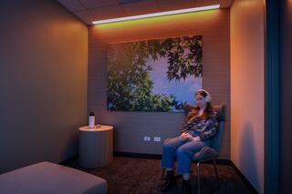 A woman sits in a softly lit corner of a quiet room with headphones on, surrounded by warm tones and a nature-themed painting, creating a calm, relaxing atmosphere.