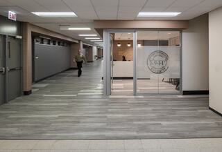 A hallway leads to a glass-fronted 911 dispatch center, with a person walking by and modern, well-lit surroundings.
