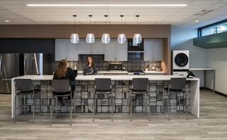 A clean, sleek new kitchen features updated cabinetry, illuminated pendant lights, and a spacious bar with two people engaged in conversation.