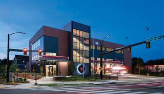 A multi-story fire station with large windows and contemporary architecture is illuminated at dusk, with fire truck leaving building.