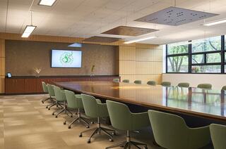 A modern conference room features a long wooden table, green chairs, a large screen, and natural light from several windows.