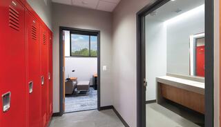 A bunk room features red lockers on one side, with a doorway leading to a room containing a bed and a bright, open bathroom.