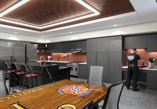 New Godfrey Fire Protection District's Station No. 1 kitchen, designed by FGM Architects; one service member works at a kitchen counter; large branded kitchen dining table is in the forefront, open kitchen with a counter-height seating and lower kitchen island, large fridges and ample cabinet space.