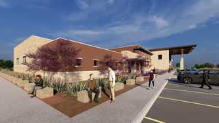 A modern building with a landscaped exterior, featuring seating areas and desert plants, under a clear blue sky. People are actively interacting outside.