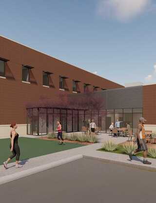 A modern outdoor area featuring a glass structure, greenery, and people walking and gathering, set against a brick building backdrop.