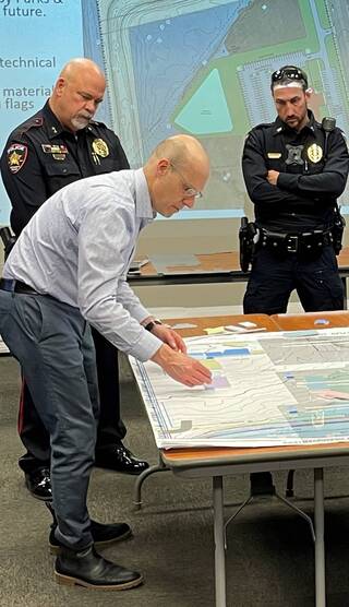 A man examines a large map at a meeting while two police officers observe, possibly discussing plans or technical details.