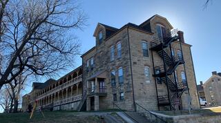 A historic stone building features a staircase, large windows, and a porch, surrounded by bare trees and clear blue skies.