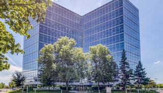 A modern glass office building with lush green trees in the foreground and a clear blue sky in the background.